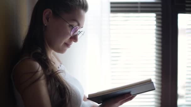 Fille avec des lunettes lit un livre près de la fenêtre avec volets, contre-jour — Video