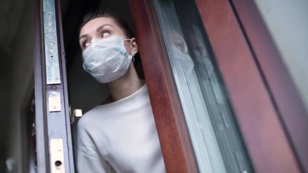 一个戴着医疗面具的女孩穿过前门的缝隙从屋里探出头来 呼吸着空气 环顾四周 关上了门 — 图库视频影像