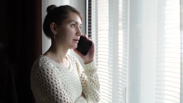 窓の近くに立って電話で話してる女の子 通りを見ている若い女性は — ストック動画
