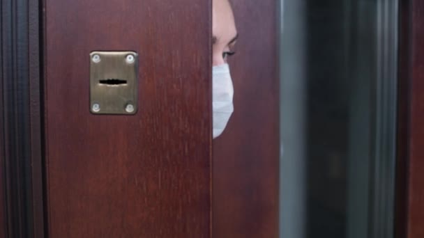 一个戴着医疗面具的女孩穿过前门的缝隙从屋里探出头来 呼吸着空气 环顾四周 关上了门 — 图库视频影像