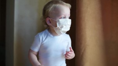 Koruyucu tıbbi maskeli küçük bir çocuk perdenin arkasından pencereden dışarı bakar, sonra perdeyi açar ve parlak güneş ışığı yüzüne düşer. Tıbbi maskeli bebek.