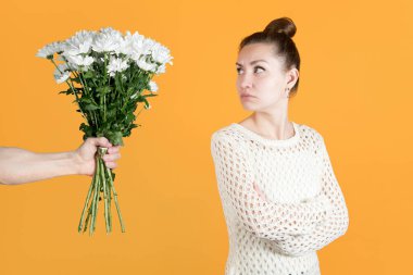 Gücenmiş kız, erkeğin ona verdiği çiçeklere omzunun üzerinden bakar. Beyaz kazaklı ve kasımpatı takmış bir kız.