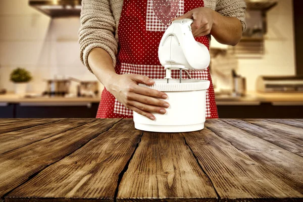 Woman in red apron with white mixer in kitchen and wooden desk top space for products and decorations.