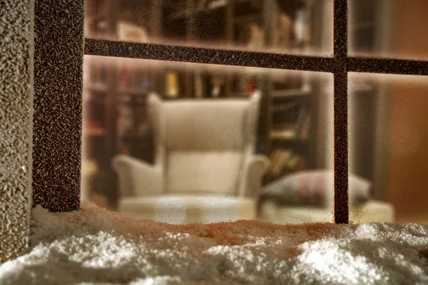 Outside snowy winter window view with cozy relaxing home interior. Looking through the window at comfortable armchair and warm atmosphere inside the room. Snow space for products and decorations.