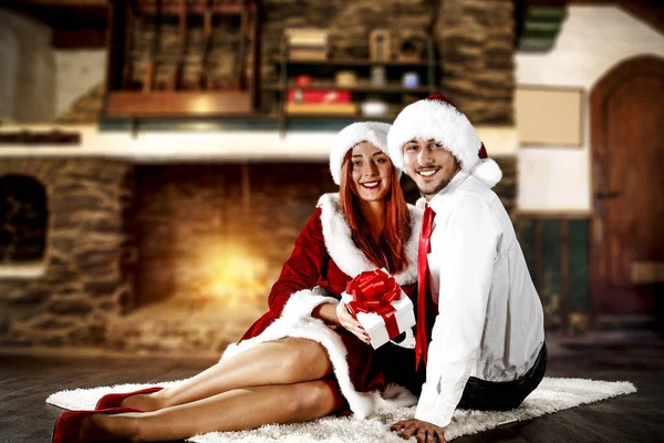 Young Santa Claus people with a present with old fireplace background. Free space for your decoration. Chrsitmas time and cold winter night. Old fireplace wall background. Woman and man smiling and lying on the white carpet at the fireplace.