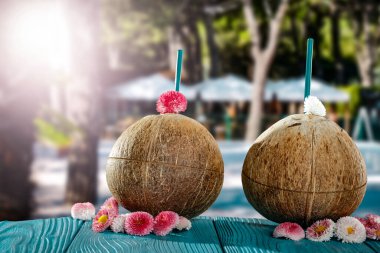 Taze hindistan cevizi içeceği, bulanık egzotik ağaçlar ve açık hava restoran geçmişi. Güzel güneşli yaz dönemi arkaplanı. Reklam ürünleri için yer kopyala. Hindistan cevizleri ve çiçekler, tahta masadaki çiçek yaprakları..