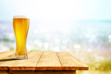 Büyük bardakta soğuk bira, ahşap masada, okyanus ve kumlu plaj arka planında. Reklam ürünleri için alanı kopyala.