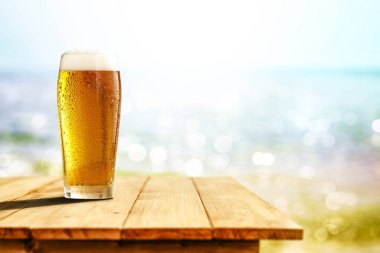 Büyük bardakta soğuk bira, ahşap masada, okyanus ve kumlu plaj arka planında. Reklam ürünleri için alanı kopyala.