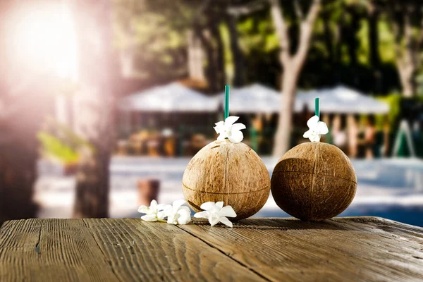 Fresh coconut drink and blurred exotic trees and outdoor restaurant background. Beautiful sunny summer time background. Copy space for advertising products. Coconuts and flowers, petals of flowers on wooden table.