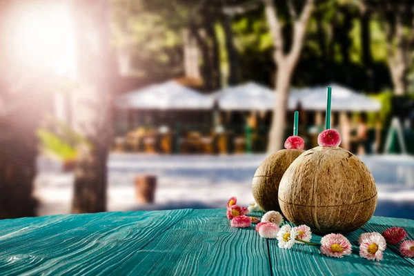 Fresh coconut drink and blurred exotic trees and outdoor restaurant background. Beautiful sunny summer time background. Copy space for advertising products. Coconuts and flowers, petals of flowers on wooden table.