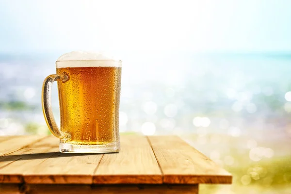 Cold beer in big glass on wooden table with ocean and sandy beach background. Copy space for advertising product.