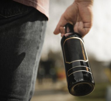 Elinde koyu bir şişe şarap, yeşil manzaralar ve üzüm bağları tutan bir adam.