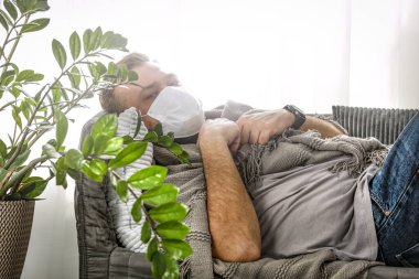 Yüzü maskeli, evde oturup dinlenen hasta bir adam. Ev karantinası. Evde büyük pencere var. Televizyon izliyordum. Güneşli bahar sabahı.