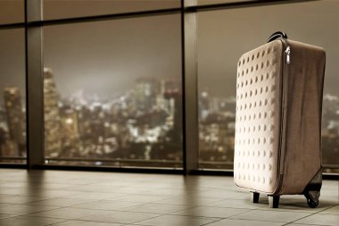 Büyük cam pencereli bir dairede yerde bavul ve geceleri bulanık bir şehir.