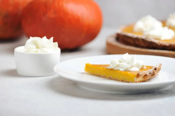 A slice of pumpkin pie tart with whipped cream.  In the background is a pie and pumpkins.  Light background.