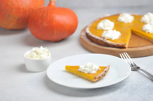 A slice of pumpkin pie tart with whipped cream.  The cake is cooked for Thanksgiving.  In the background is a pie and pumpkins.  Light background.