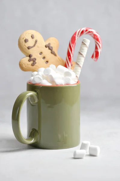 Hot drink with marshmallows. In a cup there is a gingerbread man, a candy Christmas cane and a waffle roll. Light background. The vertical orientation of the frame.