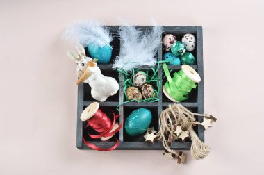 Paskalya kompozisyonu oluşturmak için eşyalarla dolu bir kutu. Ayrı bölümlerde tüyler, kurdeleler, bıldırcın yumurtaları, tavuk yumurtaları ve tavşan heykelciği bulunur. Hafif arkaplan. Metin için boş yer.