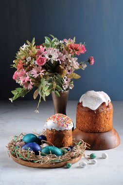 Paskalya kompozisyonu: maya hamurundan yapılmış yuva ve fırın ürünleri olarak stilize edilmiş bir tabağın üzerinde mavi ve yeşil renkli yumurtalar. Çerçevenin dikey yönelimi. Koyu arkaplan.