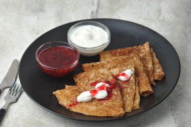 Siyah bir tabakta, ekşi krema ve reçelli ince krepler. Hafif arkaplan. Yakın plan. Çerçevede çatal bıçak takımı var..  