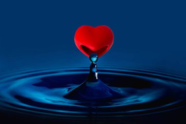 Kırmızı kalp su damlası yiyor. Cam bardağa su sıçratıyor. Mavi renk. Sevgililer Günü konsepti.