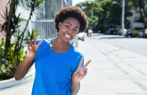 Mujer africana en una camisa azul mostrando signo de victoria al aire libre en ci —  Fotos de Stock