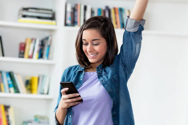 Cheering spanish young adult woman sending message with mobile phone indoors at home