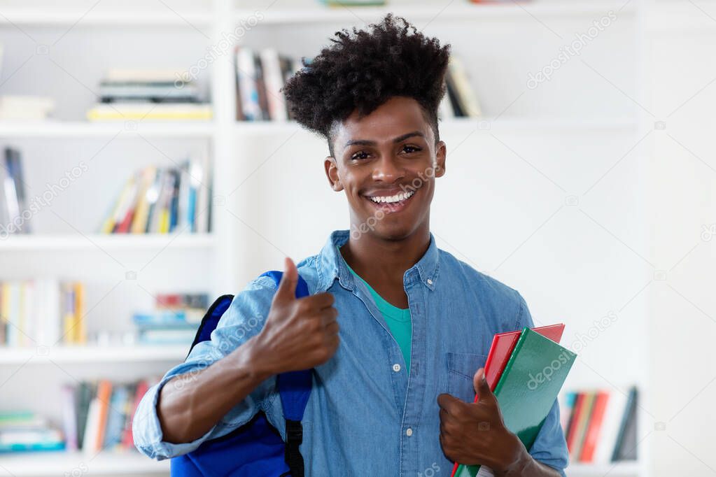 Afro american male student with backpack and paperwork showing thumb up at classroom of university
