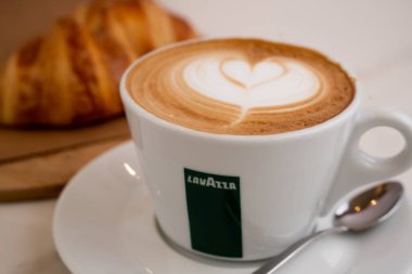 Arka planda kapuçino resmi, yanında kruvasan olan lavza bardağında.
