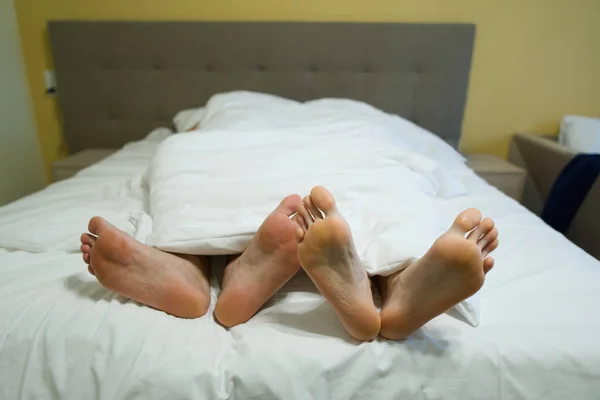 Dois pares de pernas na cama sob um cobertor branco. masculino e feminino mentira pés no vermelho cama juntos . — Fotografia de Stock