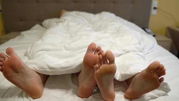 Tre paia di gambe a letto sotto una coperta bianca, giocando — Video Stock