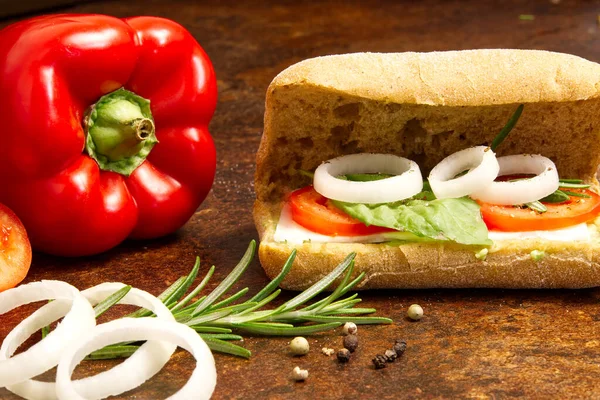 Sándwich hecho de ciabatta de pan italiano sobre una mesa marrón. cebolla, tomate, pimienta, queso de cabra, razmoin, albahaca y aguacate . — Foto de Stock