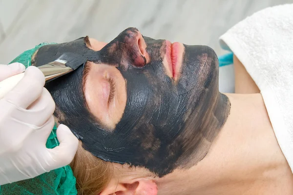 Косметик наносит натуральную увлажняющую маску на лицо молодой красивой женщины. Черный бамбук Угольная маска для лица, нанесенная на лицо кистью . — стоковое фото