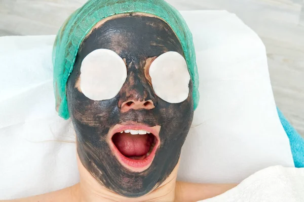 Young woman in a beauty salon with a black mask on her face. The concept of cosmetology. The cosmetologist performs a procedure to cleanse the face with problem skin. — Stock Photo, Image