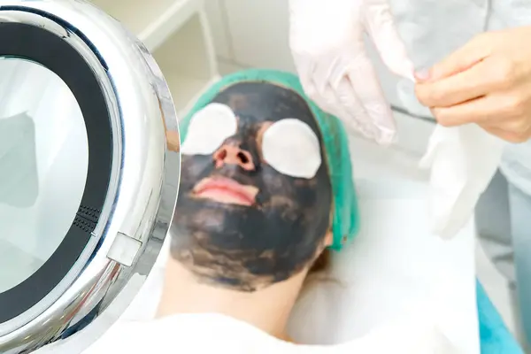 Молодая женщина в салоне красоты с черной маской на лице. Понятие косметологии. Косметолог проводит процедуру очищения лица проблемной кожей. под косметическим лупой . — стоковое фото