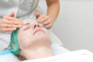 Güzellik uzmanı genç ve güzel bir kadının yüzüne doğal nemlendirici bir maske takar. Yaşlanma önleyici ve kırışıklık önleyici maske bir fırçayla yüze uygulanır. Kozmetoloji konsepti. Kozmetik uzmanı 