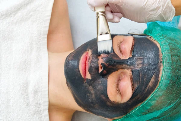 Beautician brengt natuurlijke hydraterende masker op het gezicht van een jonge mooie vrouw. Zwarte bamboe Charcoal Face Mask, aangebracht op het gezicht met een borstel. — Stockfoto