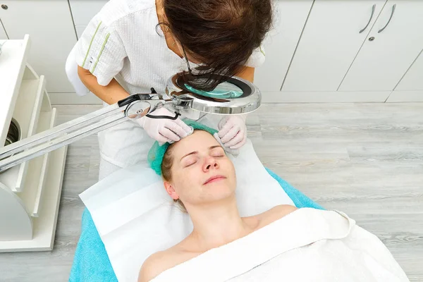 Schoonheidsspecialiste reinigt gezicht. Jonge vrouw in een schoonheidssalon. Cosmetologisch concept. De cosmetoloog voert een procedure uit om het gezicht te reinigen met een probleemhuid. onder cosmetisch vergrootglas. — Stockfoto