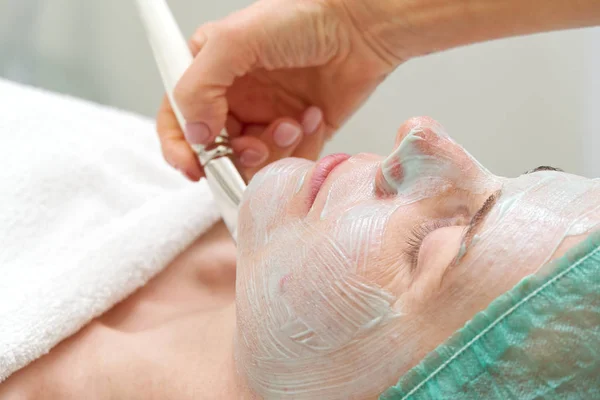 Beautician applicerar en naturlig fuktgivande mask på ansiktet av en ung vacker kvinna. Anti-aging och rynka mask appliceras på ansiktet med en borste. Kosmetologiskt koncept. Kosmetologen utför — Stockfoto