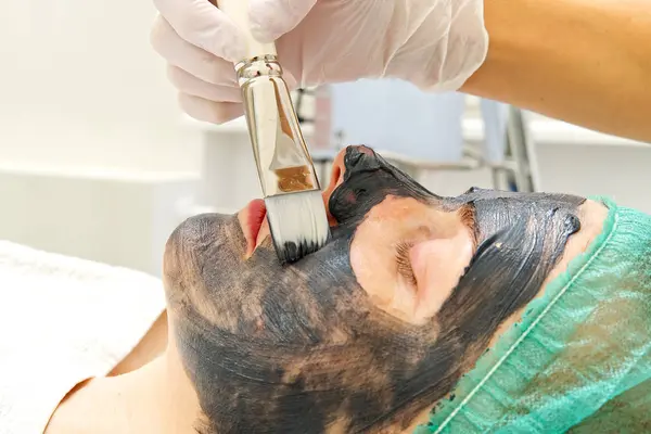 Beautician brengt natuurlijke hydraterende masker op het gezicht van een jonge mooie vrouw. Zwarte bamboe Charcoal Face Mask, aangebracht op het gezicht met een borstel. — Stockfoto