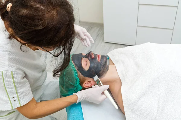 Beautician brengt natuurlijke hydraterende masker op het gezicht van een jonge mooie vrouw. Zwarte bamboe Charcoal Face Mask, aangebracht op het gezicht met een borstel. — Stockfoto