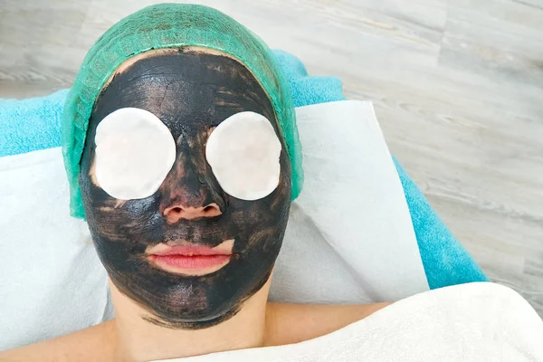 Young woman in a beauty salon with a black mask on her face. The concept of cosmetology. The cosmetologist performs a procedure to cleanse the face with problem skin.