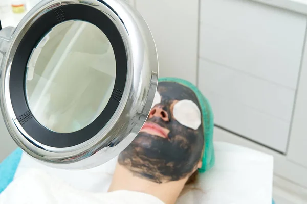 Jonge vrouw in een schoonheidssalon met een zwart masker op haar gezicht. Het concept van kosmetologie. De cosmetoloog voert een procedure uit om het gezicht te reinigen met een probleemhuid. onder cosmetisch vergrootglas. — Stockfoto