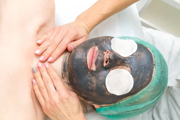 Jonge vrouw in een schoonheidssalon met een zwart masker op haar gezicht. Het concept van kosmetologie. De cosmetoloog voert een procedure uit om het gezicht te reinigen met een probleemhuid. — Stockfoto