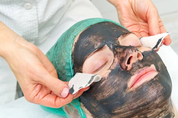 Schoonheidsspecialiste wast een natuurlijk hydraterend masker af op het gezicht van een jonge mooie vrouw. Zwart bamboe masker. Sluiten foto — Stockfoto