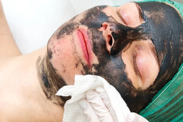 Beautician washes off a natural moisturizing mask on the face of a young beautiful woman. Black bamboo mask. Close up photo — 스톡 사진