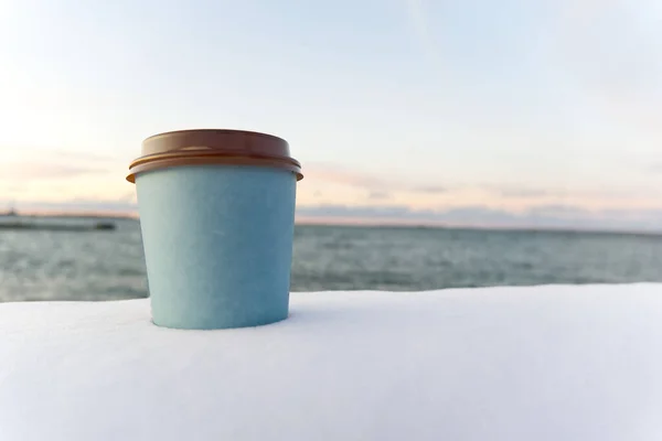 Mengambil kopi dengan latar belakang laut pada waktu musim dingin. salju musim dingin mengambil cangkir kopi matahari terbenam di laut — Stok Foto