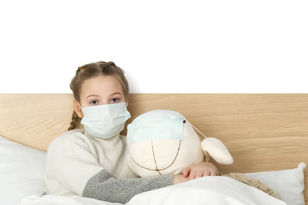 Ένα άρρωστο θλιμμένο παιδί με μάσκα με πυρετό και πονοκέφαλο ξαπλώνει στο κρεβάτι. Το βακτήριο του ιού της γρίπης κρυολογεί. κορίτσι με παιχνίδι στο δωμάτιο κρεβάτι φορούν μάσκα προστασίας, Protect Against Coronavirus έννοια — Φωτογραφία Αρχείου