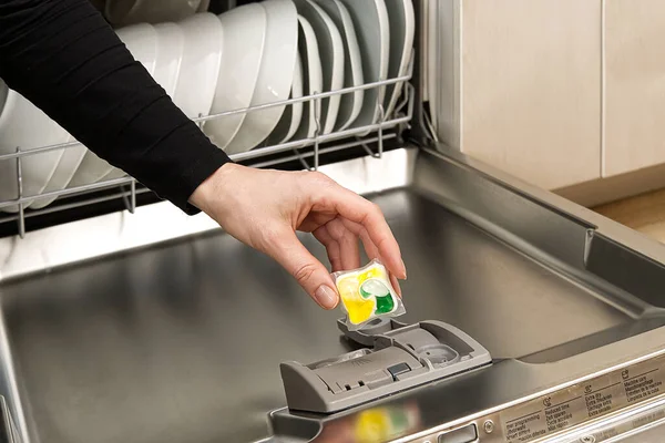 Putting tab into full integrated dishwasher close up. dishwasher machine full loaded. woman hand holding dishwasher detergent tablet.