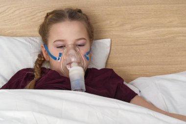 Astım maskeli kız, astım için solunum problemleri. Maske spreyli bir kız yatakta bir oyuncakla yatıyor ve adrenalin soluyor. Sağlık ve hasta çocuk konsepti